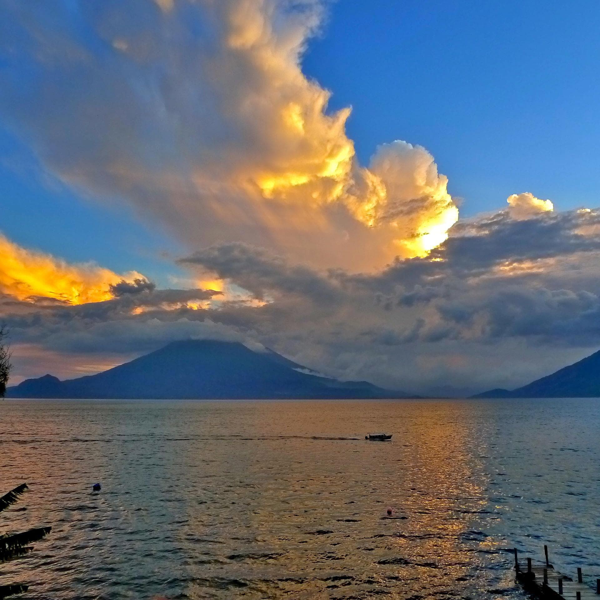 Storm light breaking gold over the volcanoes at sunset, a lone boat on Lake Atitlán